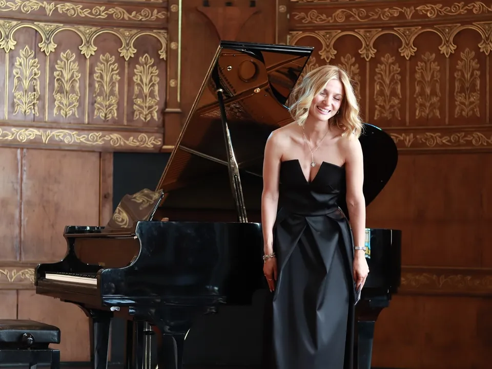 Melis Mazmanoğlu, konser salonunda kuyruklu piyanonun yanında ayakta, içtenlikle gülüyor